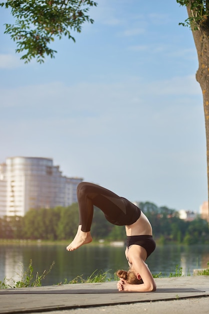 Young woman instructor doing yoga near lake in city park on summer sunny morning