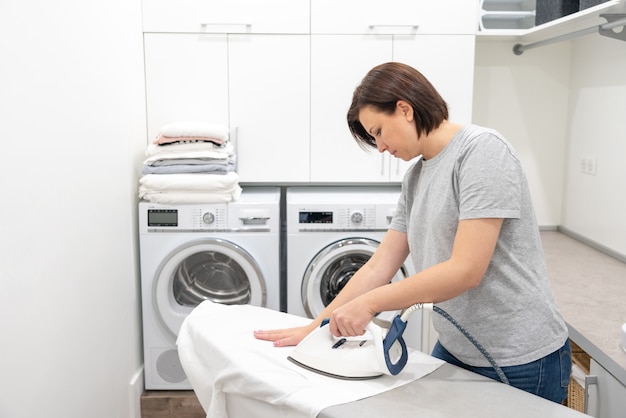 Young Woman ironing white shirt on board in laundry room with washing machine 