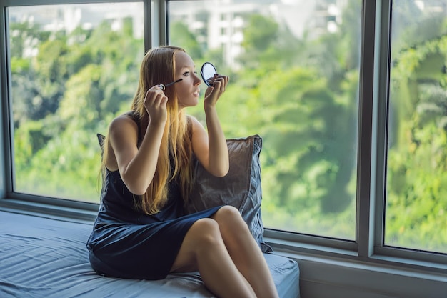 Young woman is applying makeup front of the window in beauty studio apartment Professional makeup hair styling Skin tone Bright color make up with long lashes and fresh young skin