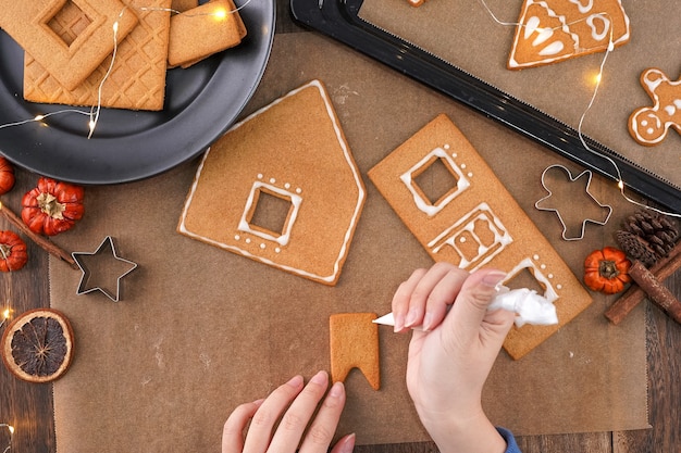 Photo young woman is decorating christmas gingerbread house cookies biscuit at home with frosting topping in icing bag, close up, lifestyle.