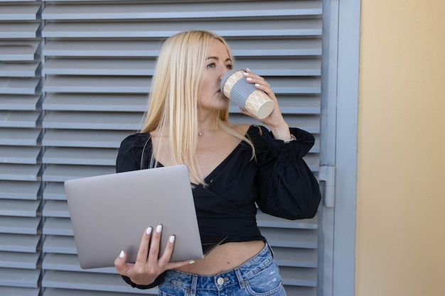 A young woman is holding a laptop and a coffee cup while working outside