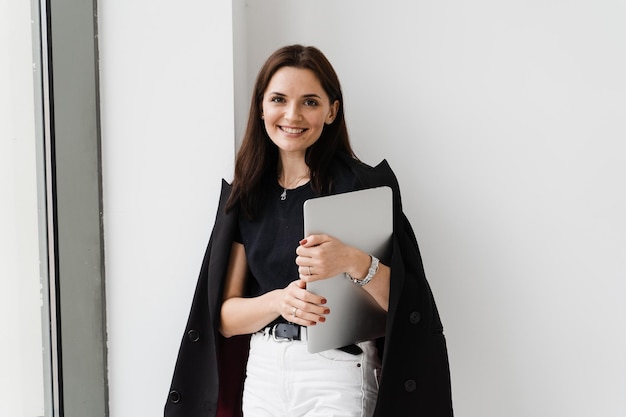 Young woman is hugging her laptop and smiling in cafe Cheerful girl with laptop standing near big window in the white office