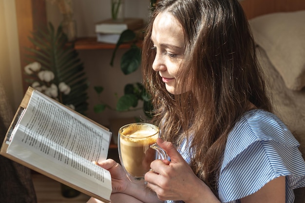 Young woman is reading a book and drinking coffee sitting by the window Concept of morning enjoyment leisure pleasant pastime and relaxation