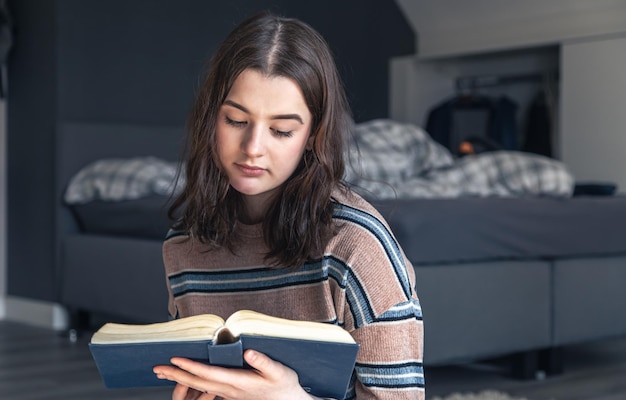 A young woman is reading a book while sitting on the floor in her room