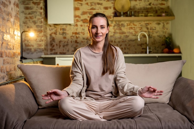 Young woman is resting at home meditating with her eyes closed sitting on the sofa in the living room a brunette housewife in the lotus position smiles