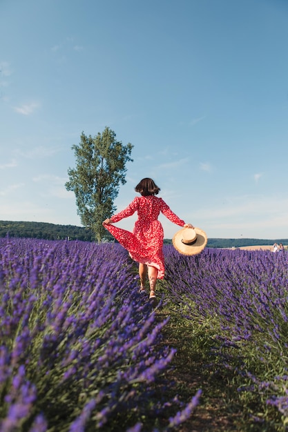 A young woman is running in a lavender field and holding a straw hat