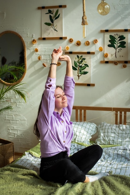 Young woman is sitting on the bed the girl stretches enjoys the rest morning in a bright bedroom