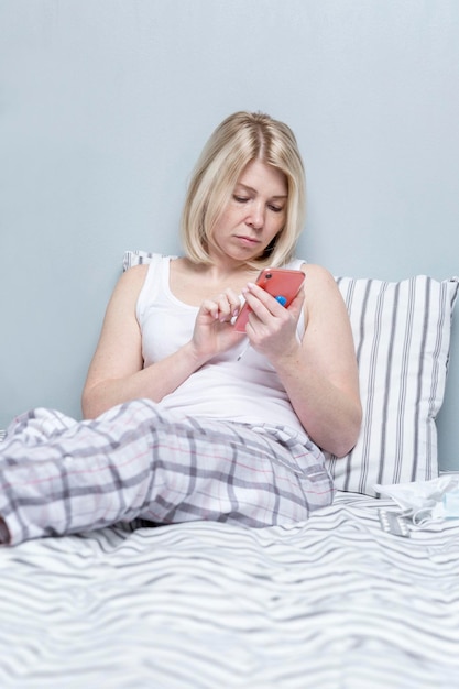 A young woman is sitting in bed with a thermometer and a phone Unpleasant symptoms of the disease and a doctor's consultation Blonde in pajamas against a gray wall Vertical