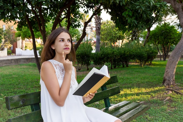 Young woman is sitting on the bench in the park and thinking about what she has read