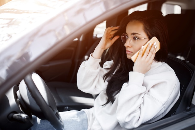 Young woman is sitting in a car. Beautiful brunette model is using a smartphone or phone