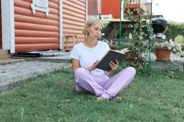 A young woman is sitting on the grass in home yard on a sunny day and reading a real book. Grass and dandilion background, nice vintage look.