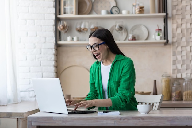 A young woman is sitting at home in the kitchen using a laptop plays online games lottery raffle