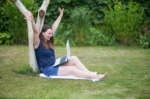 a young woman is sitting in a meadow with a laptop and talking on an online conference at work