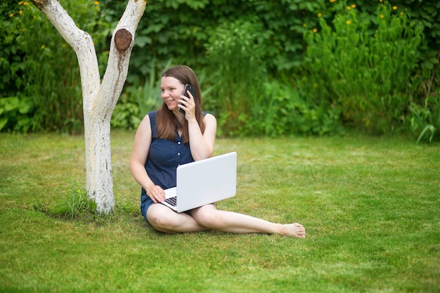 a young woman is sitting in a meadow with a laptop and talking on an online conference at work