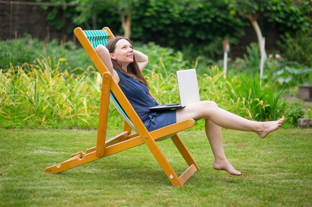 a young woman is sitting in a meadow with a laptop and talking on an online conference at work