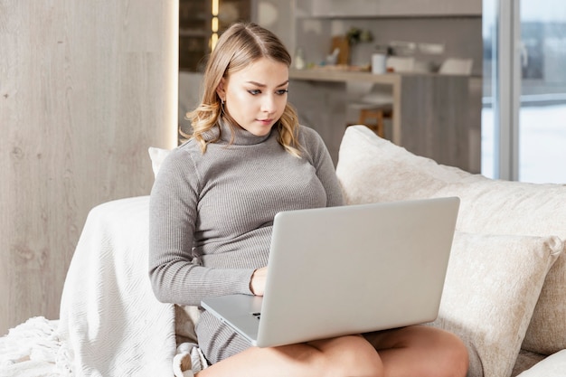 A young woman is sitting with a laptop on the couch in the living room. Beautiful blonde in a gray dress. Remote work, freelance and blogging.