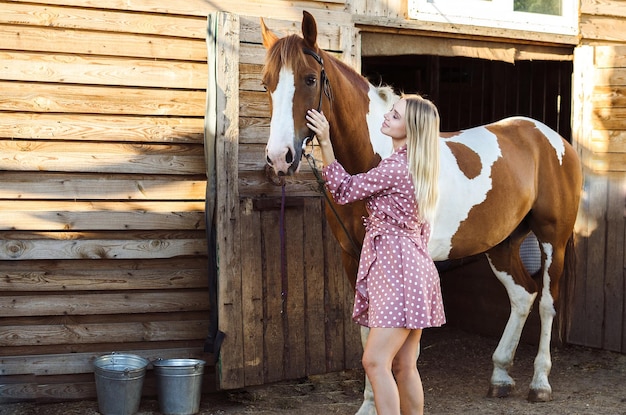 A young woman is stroking and smiling her horse in stable on a farm countryside