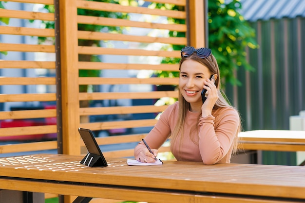 A young woman is talking on a mobile phone with a tablet and a notebook on a table in a cafe