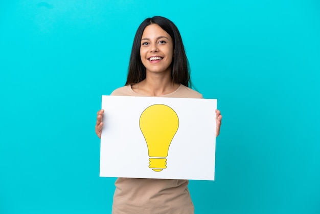 Young woman over isolated background holding a placard with bulb icon with happy expression