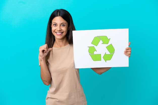 Young woman over isolated background holding a placard with recycle icon with proud gesture