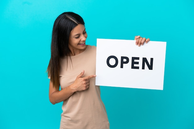 Young woman over isolated background holding a placard with text OPEN and  pointing it