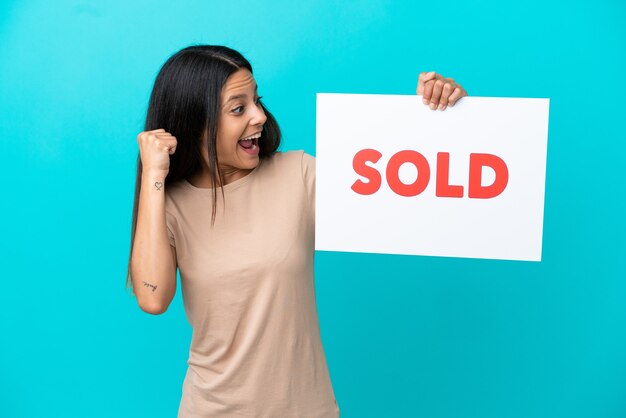 Young woman over isolated background holding a placard with text SOLD and celebrating a victory