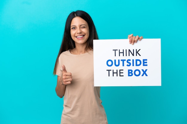 Young woman over isolated background holding a placard with text Think Outside The Box and pointing to the front