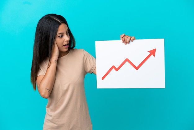 Young woman over isolated background holding a sign with a growing statistics arrow symbol with surprised expression