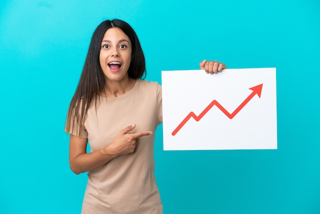 Young woman over isolated background holding a sign with a growing statistics arrow symbol with surprised expression