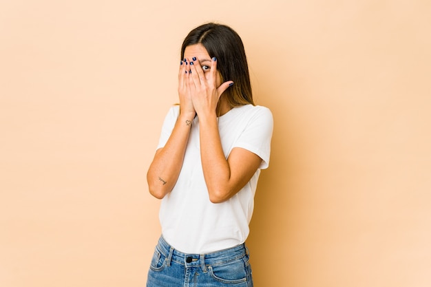 Young woman isolated on beige blink through fingers frightened and nervous.