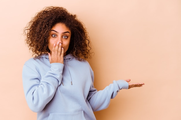 Young woman isolated on beige wall impressed holding copy space on palm