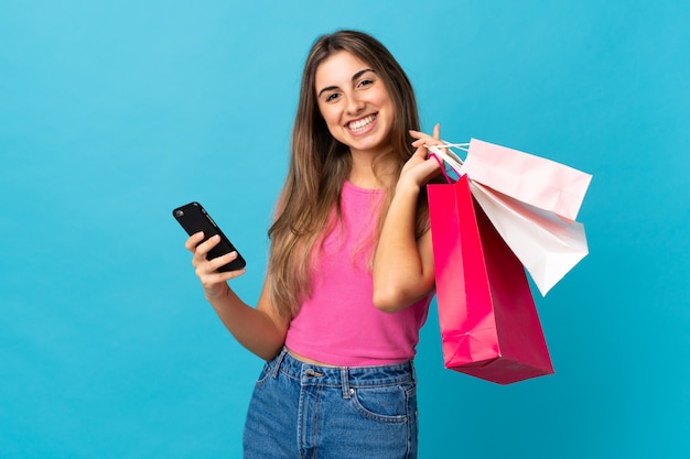 Young woman over isolated blue background holding shopping bags and writing a message with her cell phone to a friend