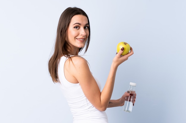 Young woman over isolated blue wall with an apple and with a bottle of water