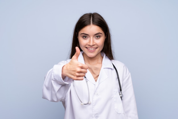 Young woman over isolated blue wall with doctor gown and with thumb up