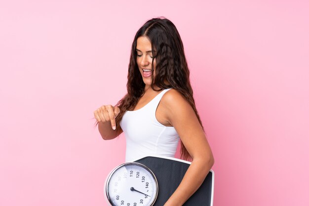 Young woman over isolated  holding a weighing machine and pointing it