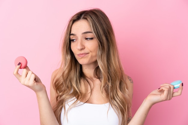 Young woman over isolated pink wall holding colorful French macarons and unhappy