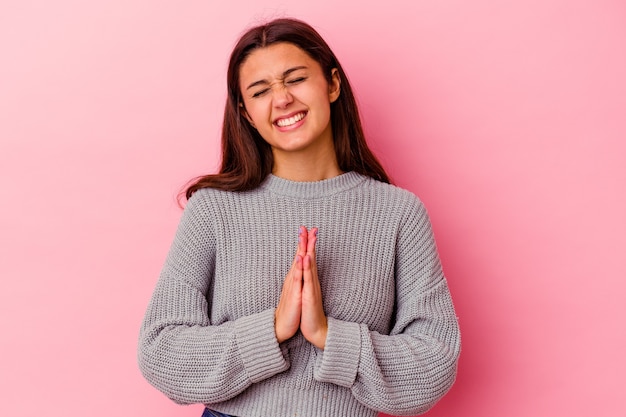 Young woman isolated on pink wall holding hands in pray near mouth, feels confident