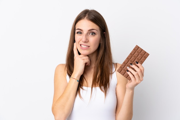 Young woman over isolated white wall taking a chocolate tablet and having doubts