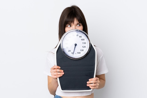 Young woman isolated on white with weighing machine and hiding behind it