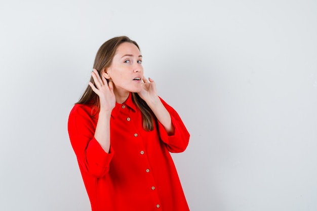 Young woman keeping hands behind ears in red blouse and looking curious , front view.