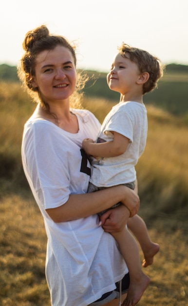 Young woman kisses her little son at sunny in the meadow in sunset. Young mother holding her baby. Mother and  little son having good time in the nature.