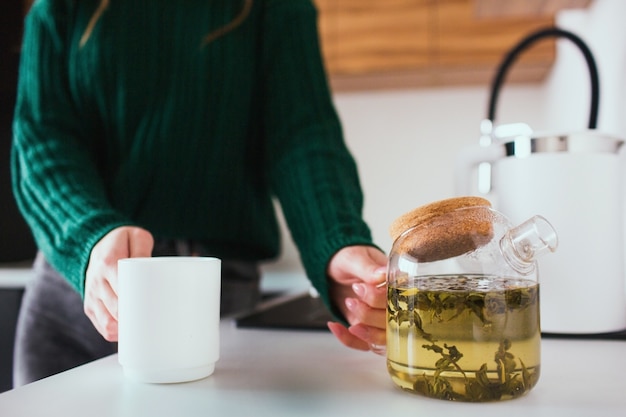 Photo young woman in kitchen during quarantine. girl hold teapot and white cup in hands. going to pour some tea for lunch or dinner.