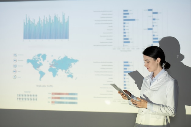 Young woman in lab coat checking information on tablet while presenting ecological project at conference