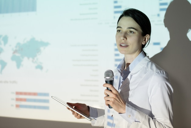 Young woman in lab coat using thesis plan on tablet while presenting project at medical convention