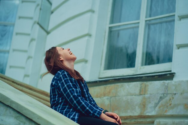 Young woman laughs sitting on steps of mansion Side view of pretty smiling female sitting near window