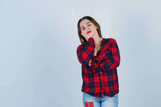 Young woman leaning cheek on palm in checked shirt, jeans and looking cheery. front view.