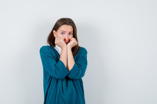 Young woman leaning cheeks on hands in sweater over white shirt and looking frightened. front view.