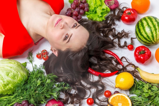 A young woman lies in fruits and vegetables on the floor on a white background Pretty serious brunette in a red dress Vegetarianism and healthy eating