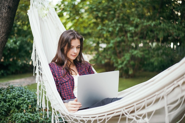 A young woman lies in a hammock with a laptop in the garden and works remotely