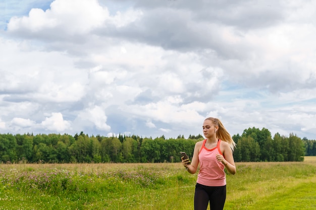 Young woman listening to music or reads messages while jogging along a path through a meadow